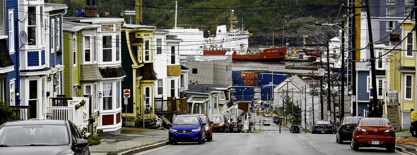 Colourful row houses line a steep street in Buckmaster's Circle, descending toward the harbour with cranes, ships and parked cars in view